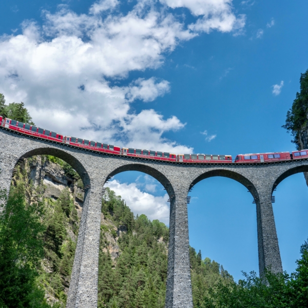 Red train crosses the iconic Landwasser Viaduct in Switzerland, surrounded by stunning alpine scenery.